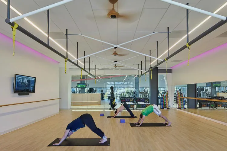 Troje ljudi u fitnes studiju izvodi joga poze na prostirkama, sa zidovima sa ogledalima i jakim osvetljenjem.