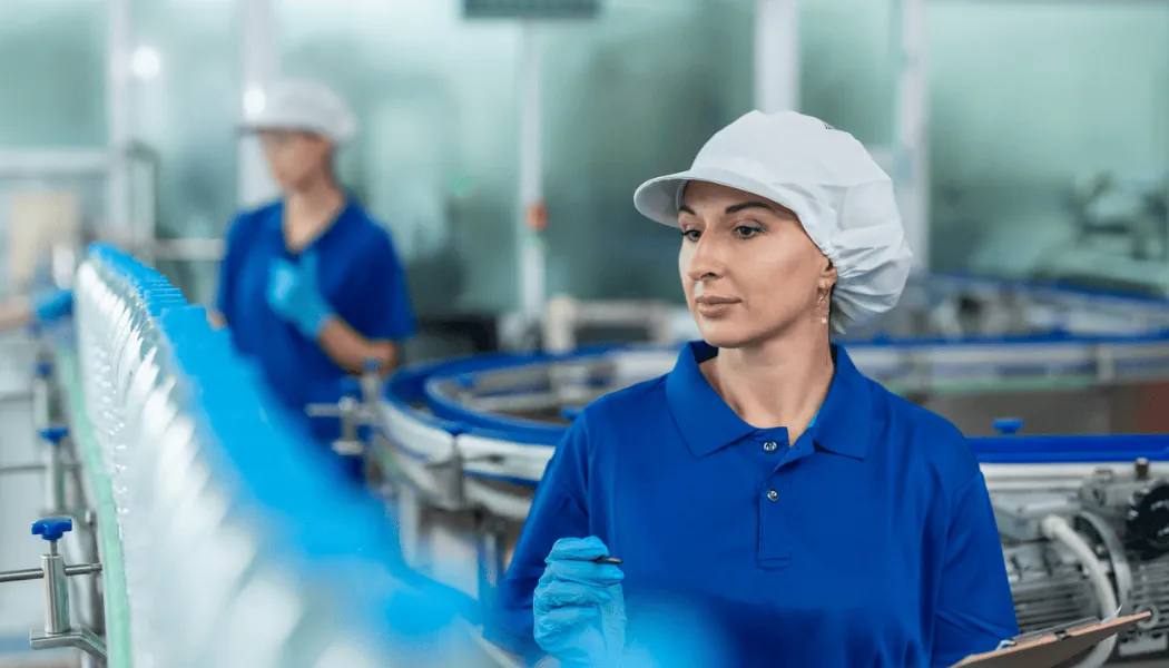 Žena u plavoj uniformi i mrežici za kosu pregleda flaširanu vodu na proizvodnoj liniji u fabrici.