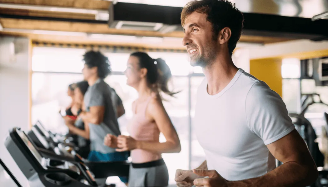 Grupa zaposlenih trči na trakama u svetlom korporativnom fitnes centru, nasmejani i usredsređeni tokom treninga.